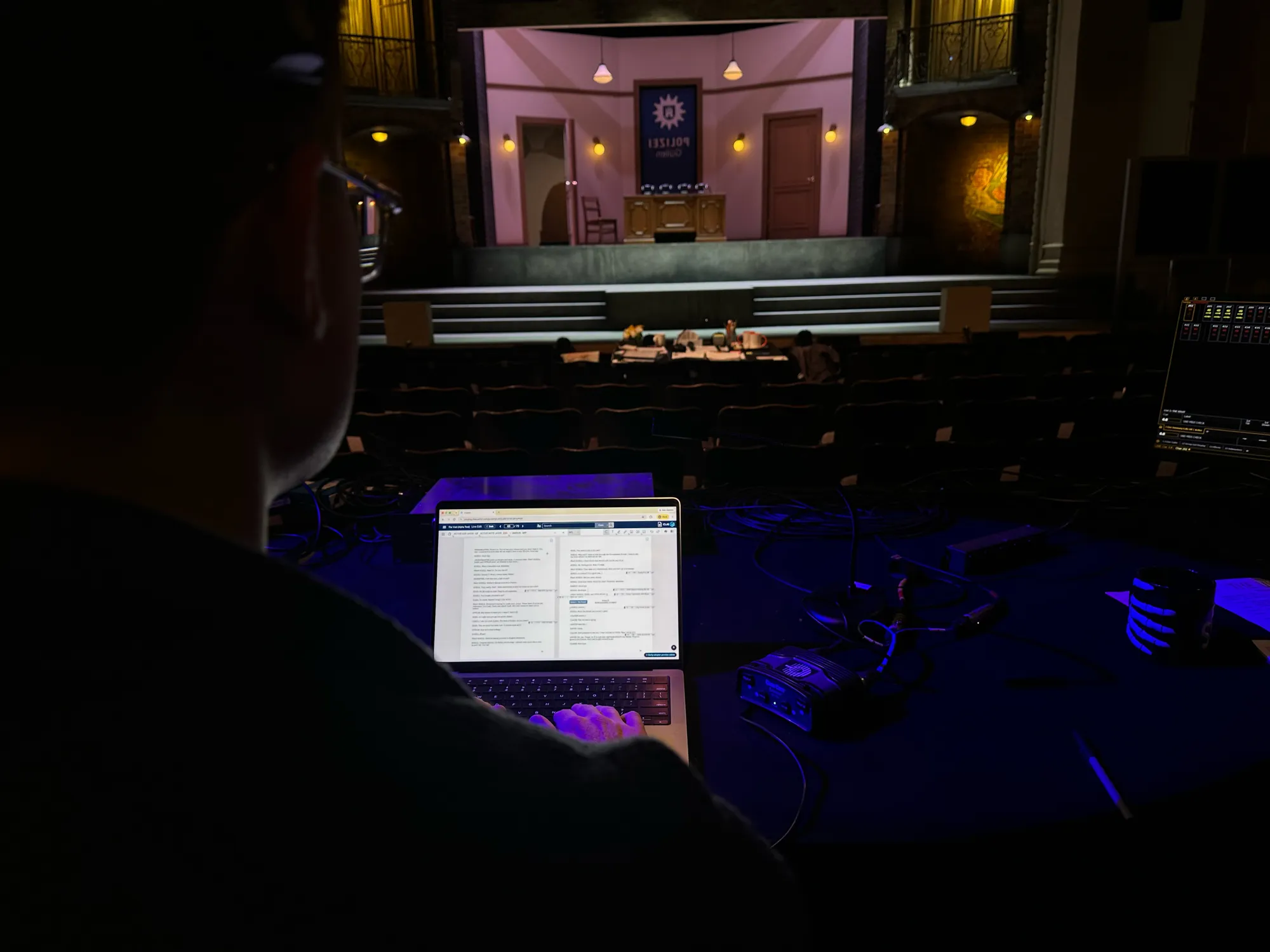 A stage manager at the calling desk during tech, headset on, running the show from Cuelist on a laptop in two-page view, with the lit set and the production table visible beyond